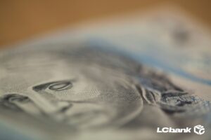 Detalhe ampliado de uma nota de real, destacando o rosto em foco suave, simbolizando dinheiro, pagamento e recebimento de valores, com logo do LCbank.